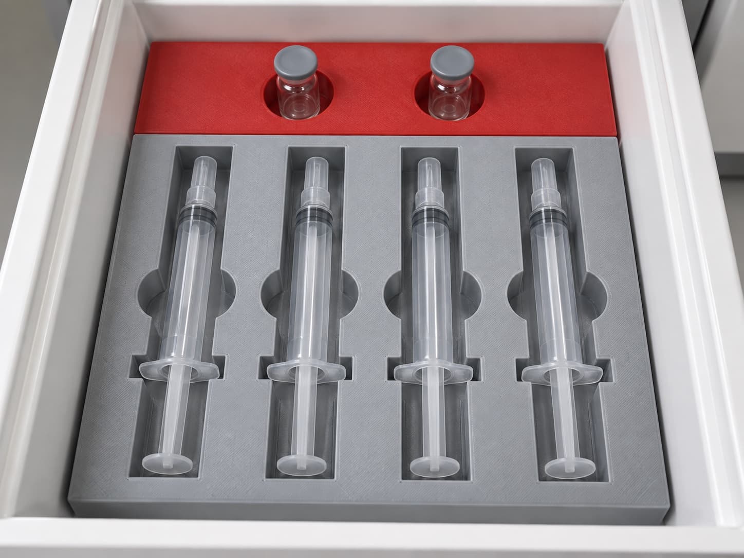 3D printed fitted drawer organizer holding four syringes in grey cradles and two vials in a red insert, in a lab cabinet drawer