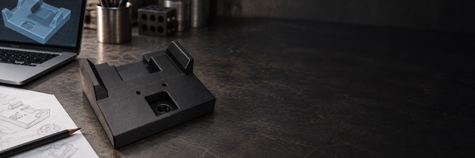 A black 3D printed locating fixture on a dark workbench, beside the hand sketch it started as and a laptop showing its CAD model — the sketch-to-part path TAKT 3D's design service runs