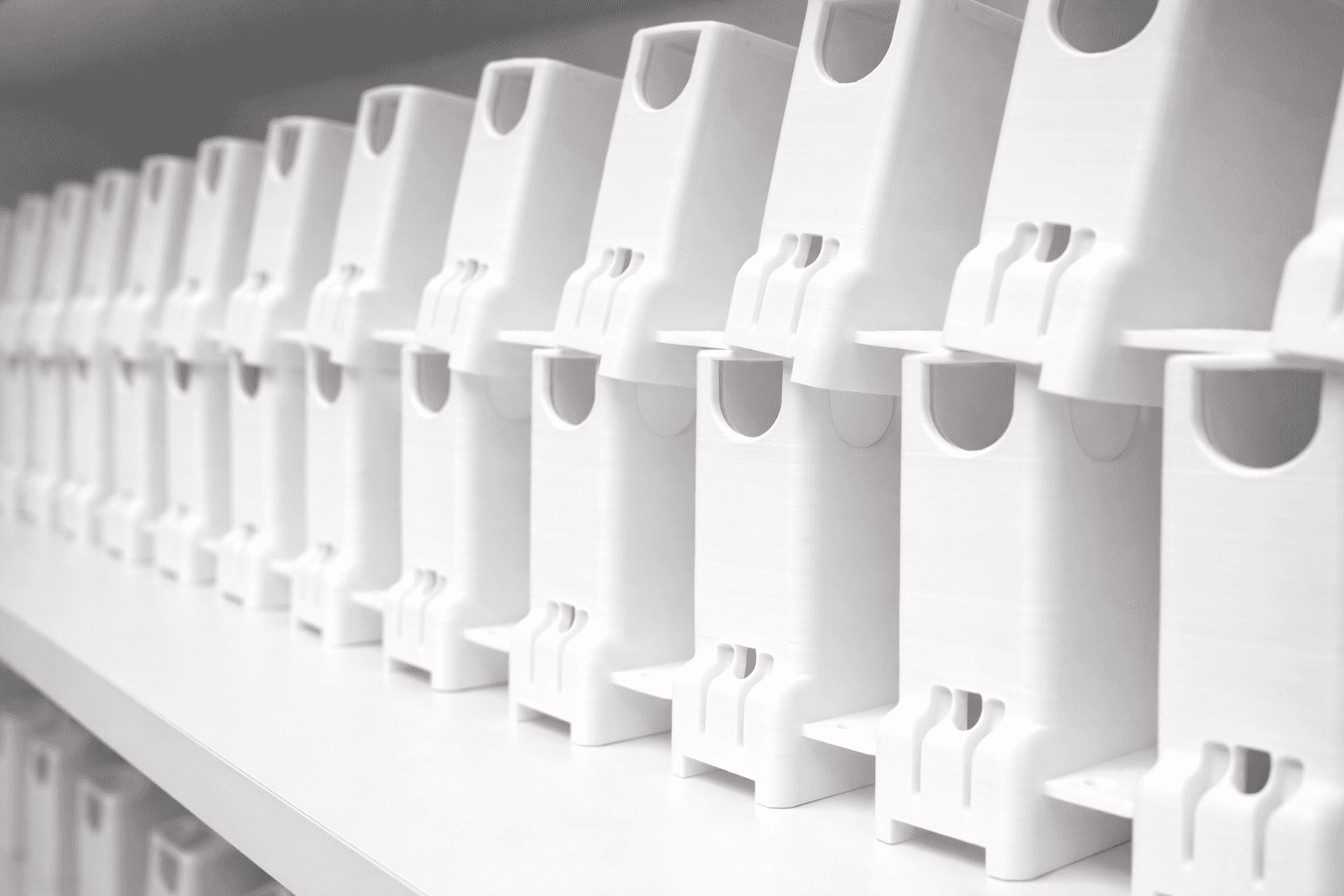 A long row of identical white 3D printed parts lined up on a shelf — a production run coming off the TAKT 3D print farm without injection tooling