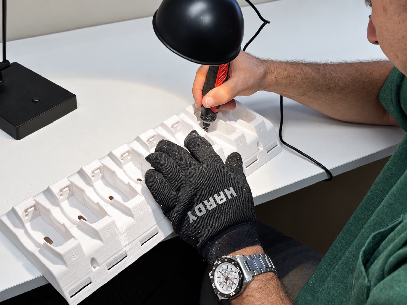A technician at a bench under a lamp, finishing a long white 3D printed fixture rail with a rotary tool