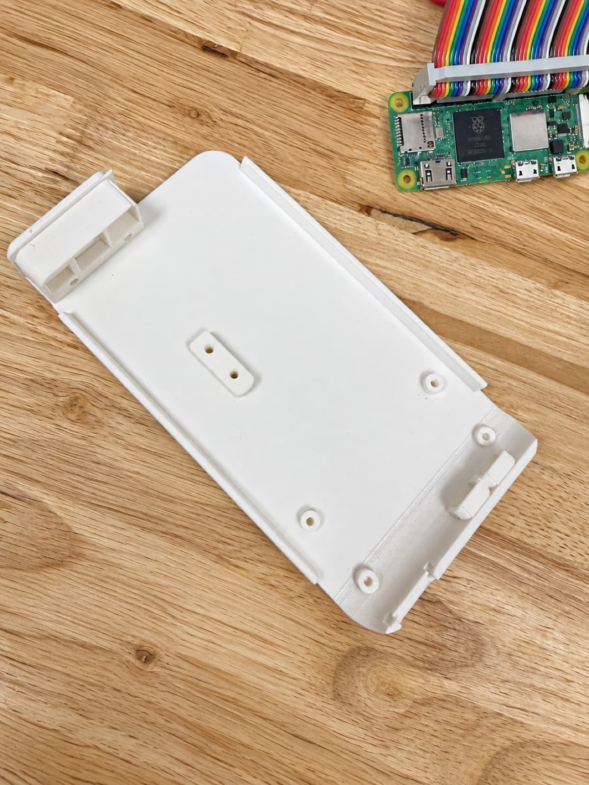 A white 3D printed electronics mounting tray with screw bosses on a wooden bench, a single-board computer and ribbon cable beside it