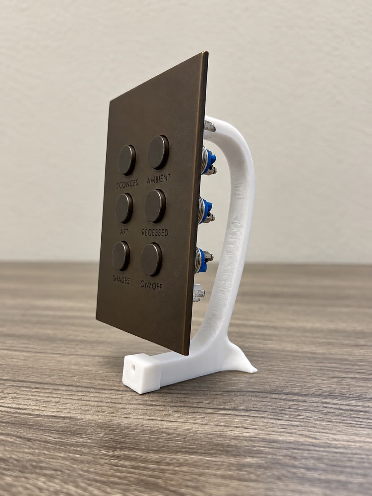A white 3D printed display stand holding a bronze six-button lighting keypad upright on a desk